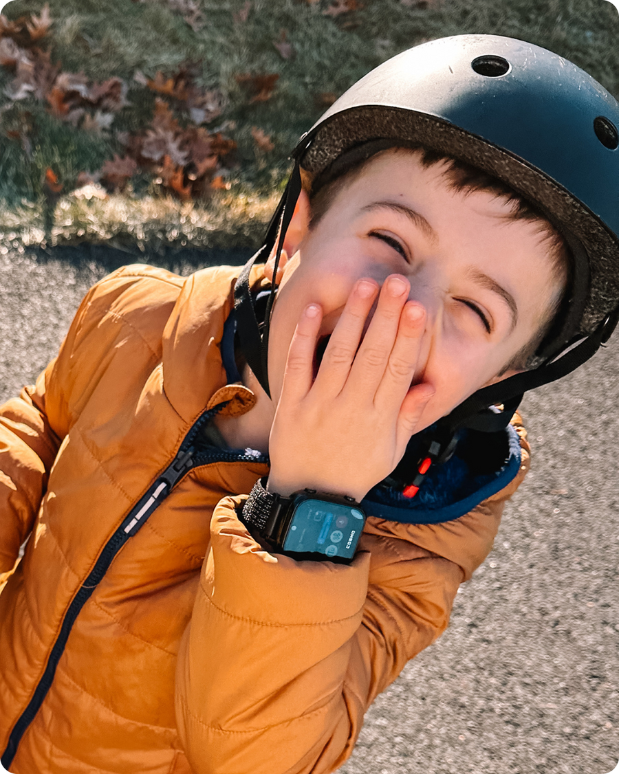 A young boy wearing a helmet laughing with his hand over his mouth, wearing a smartwatch.